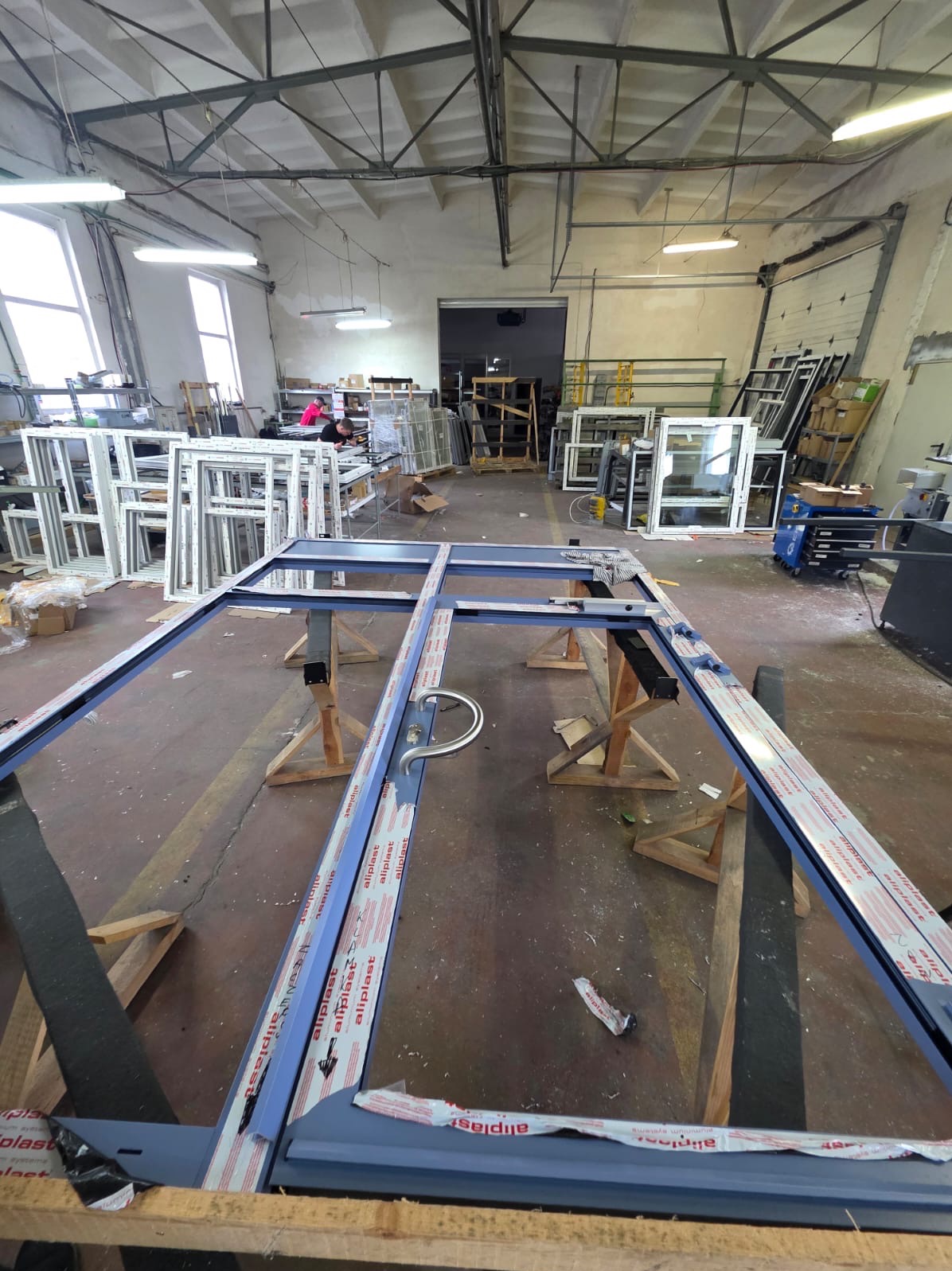 W hali produkcyjnej rama drzwiowa z szarych profili Aluplast na drewnianych kozłach. W tle okna i ramy okienne, pracownicy. Oświetlenie górne, surowy wystrój.