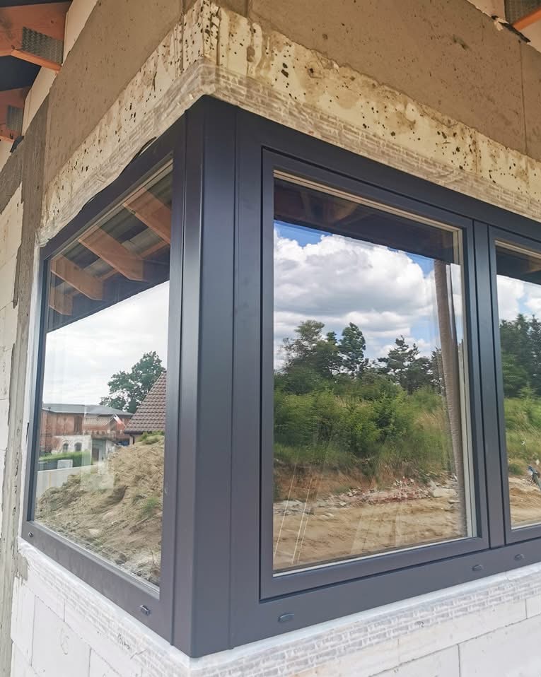 Szare okno narożne z widokiem na zewnątrz, osadzone w surowej ścianie budynku. Widoczne detale montażowe i odbicie krajobrazu w szybach.