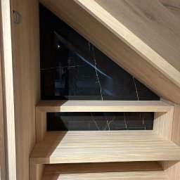 Formedge - Drewniana zabudowa sauny pod skosem dachu, z czarną płytą imitującą marmur w tle. Jasne drewno, poziome deski, dwie półki. Widoczny fragment instalacji elektrycznej.