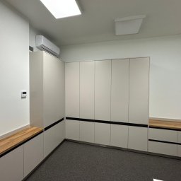 Formedge - Jasne szafy wnękowe z drewnianymi blatami w minimalistycznym wnętrzu. Czarne akcenty podkreślają nowoczesny design. Widoczny klimatyzator i oświetlenie sufitowe.