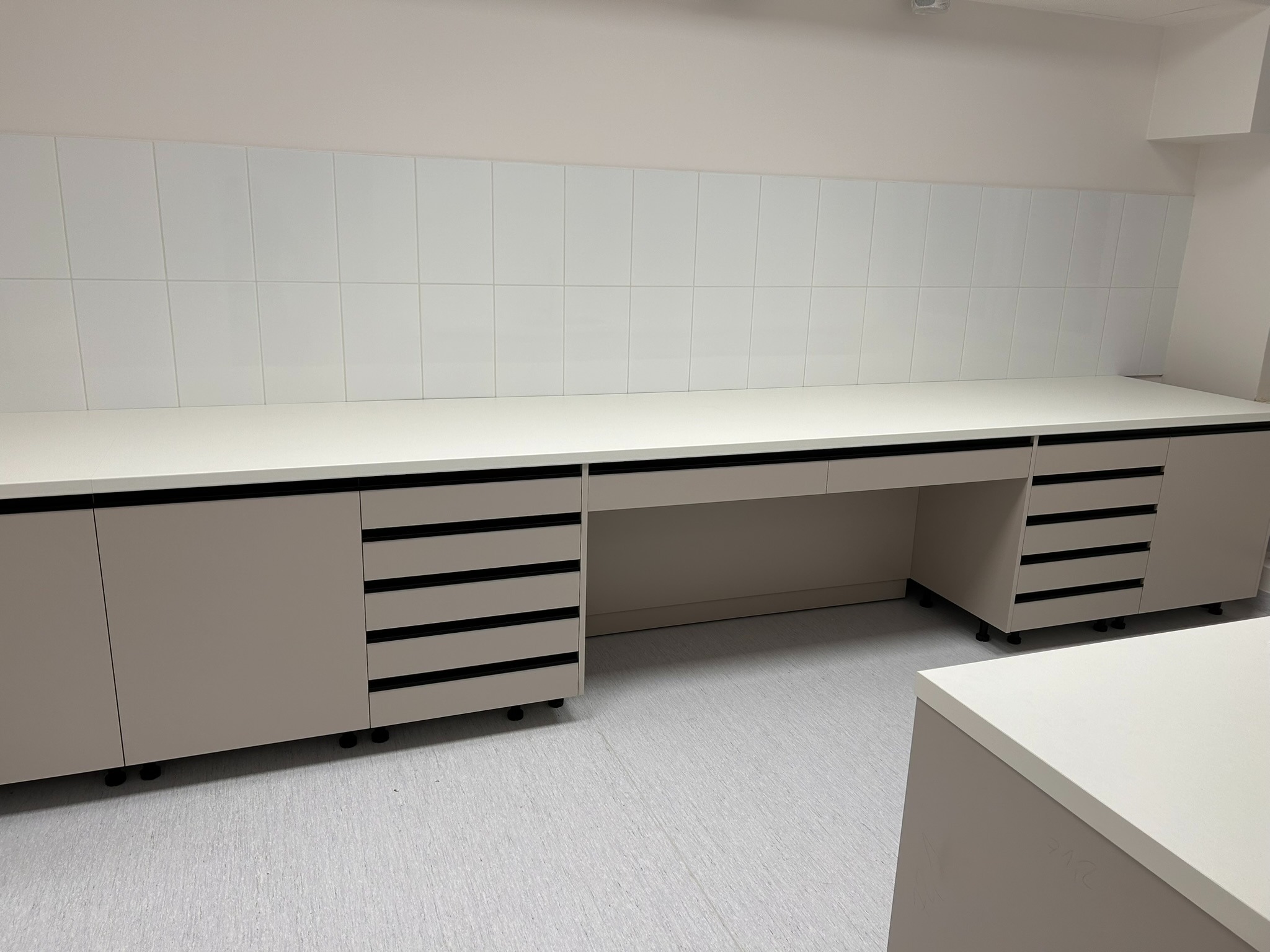 Długi, beżowy blat z szafkami i szufladami na kółkach, z czarnymi uchwytami, pod białą ścianą z płytek. Minimalistyczny design, oświetlenie sufitowe.