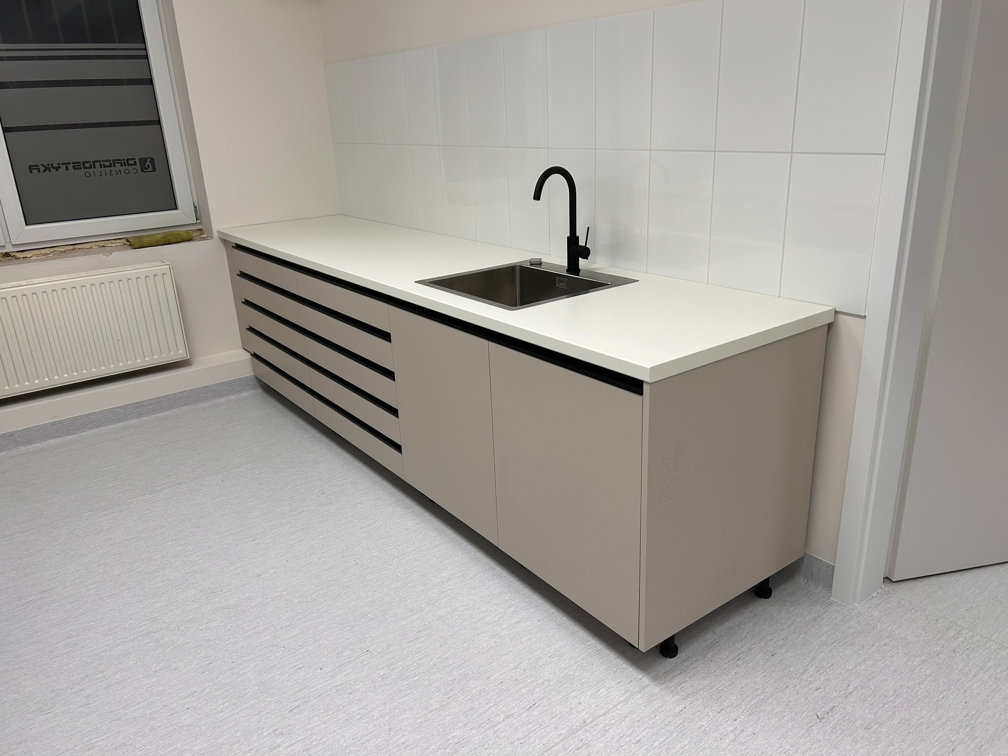 Nowoczesna zabudowa kuchenna z beżowymi frontami, białym blatem i czarną armaturą. Minimalistyczny design, płytki na ścianie, widok z perspektywy.