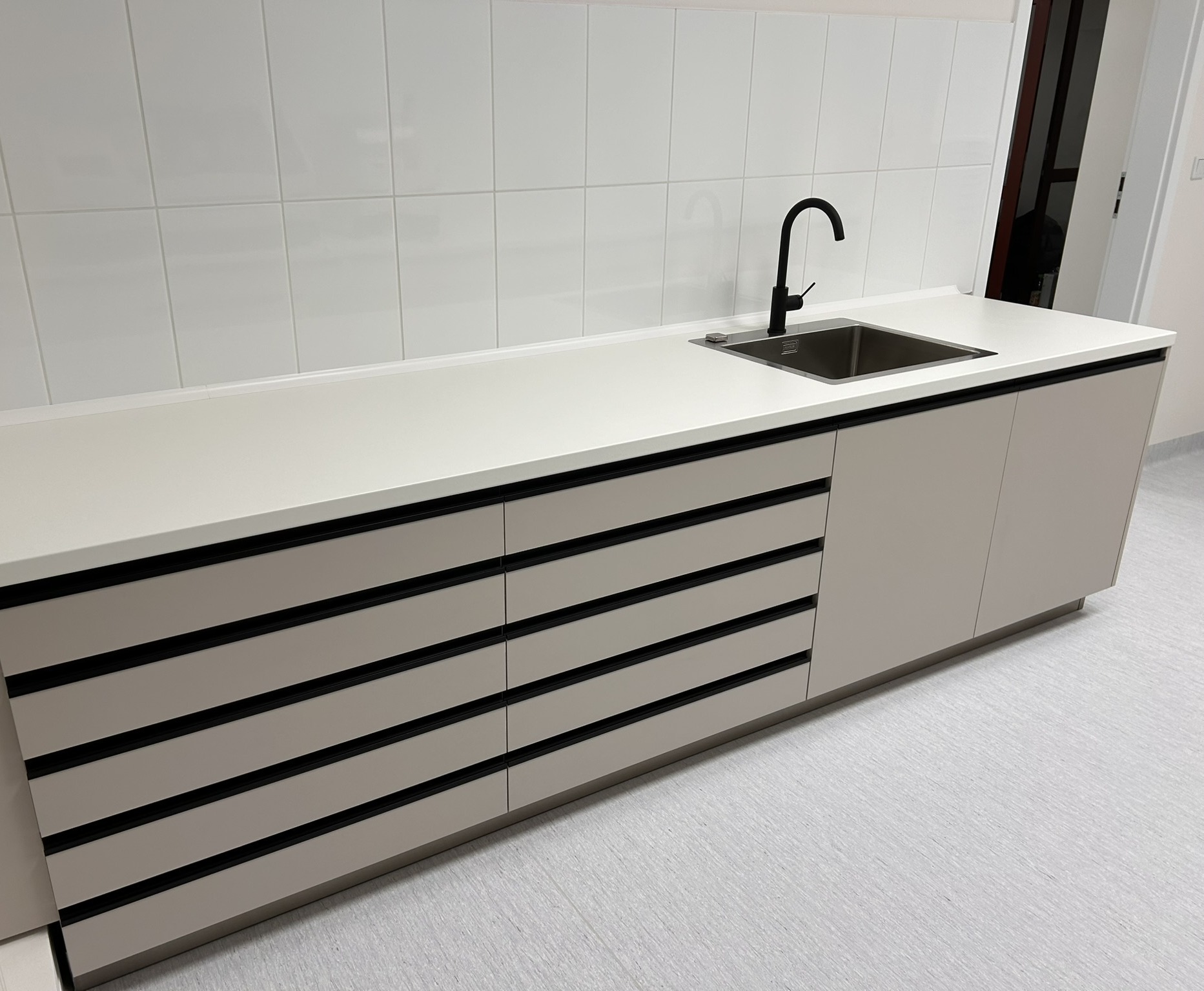 Minimalistyczna zabudowa kuchenna z białym blatem i frontami w odcieniach beżu, z czarnymi, poziomymi uchwytami. Bateria kuchenna w kolorze czarnym. Białe płytki na ścianie.