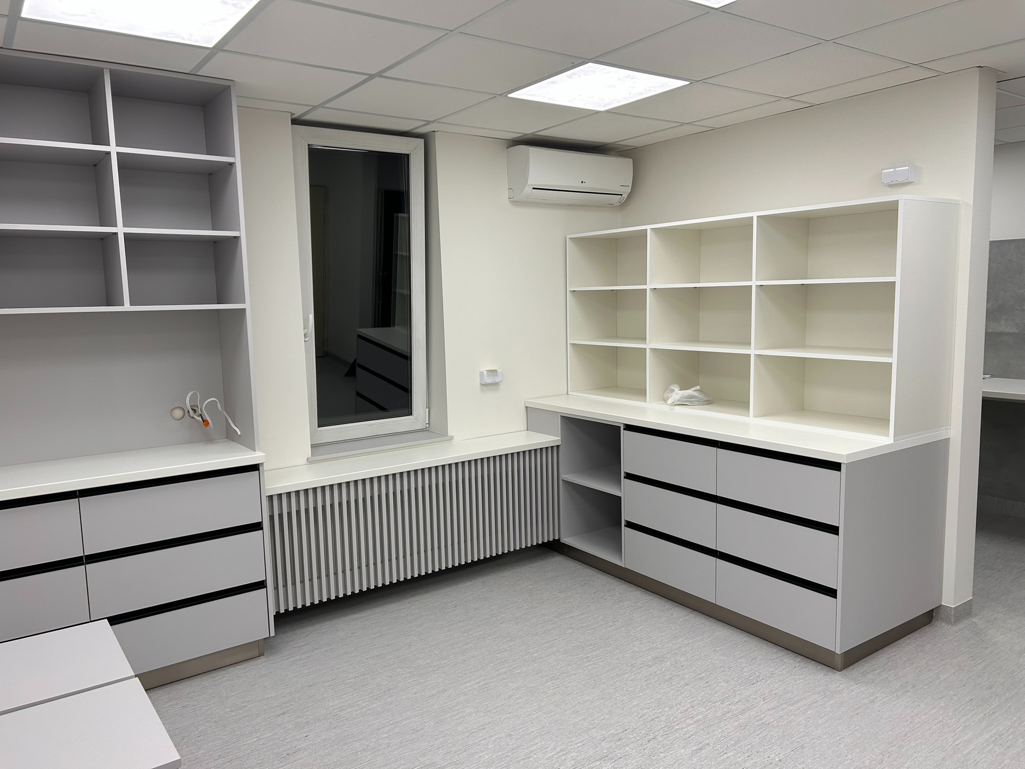 Jasne, minimalistyczne meble z szufladami i półkami w nowoczesnym wnętrzu. Widoczne detale wykończenia i oświetlenie sufitowe.