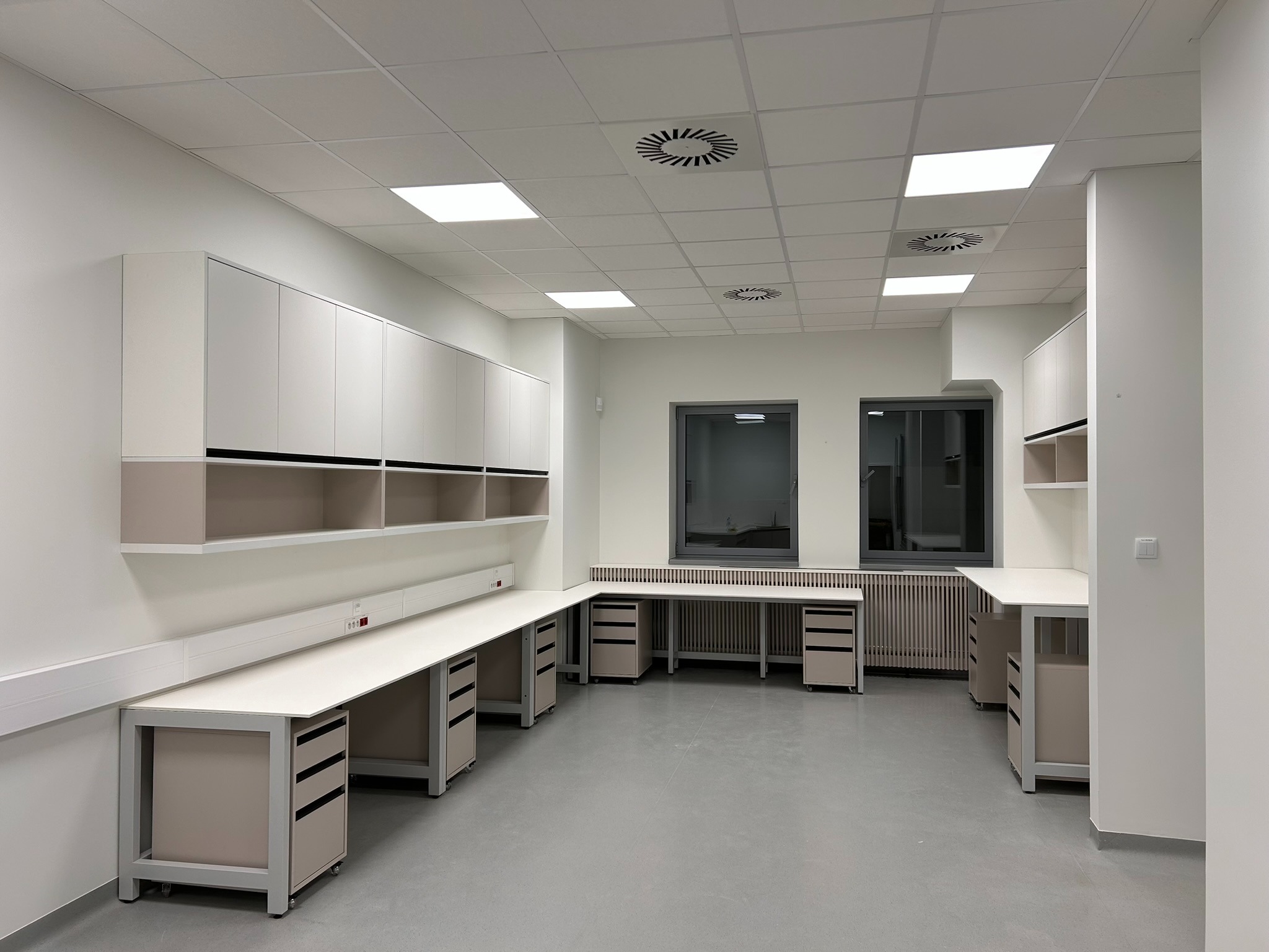 Jasne wnętrze z zabudową meblową: szafki wiszące i długi blat z szafkami na kółkach. Minimalistyczny design, neutralna kolorystyka, oświetlenie sufitowe.