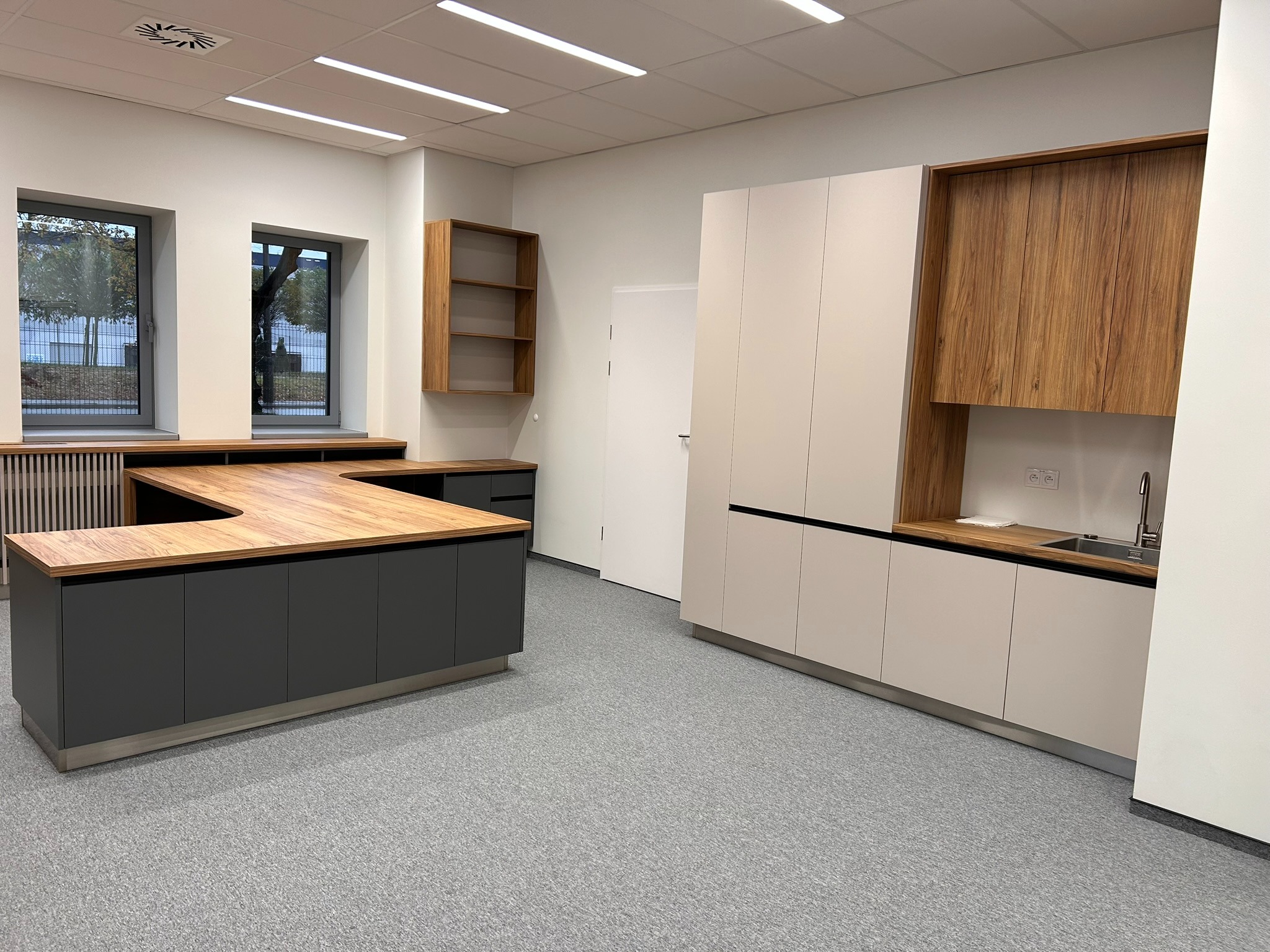 Nowoczesne meble biurowe z drewnianym blatem i szarymi szafkami, uzupełnione o zabudowę kuchenną z drewnianymi akcentami i białymi frontami, w jasnym pomieszczeniu biurowym.