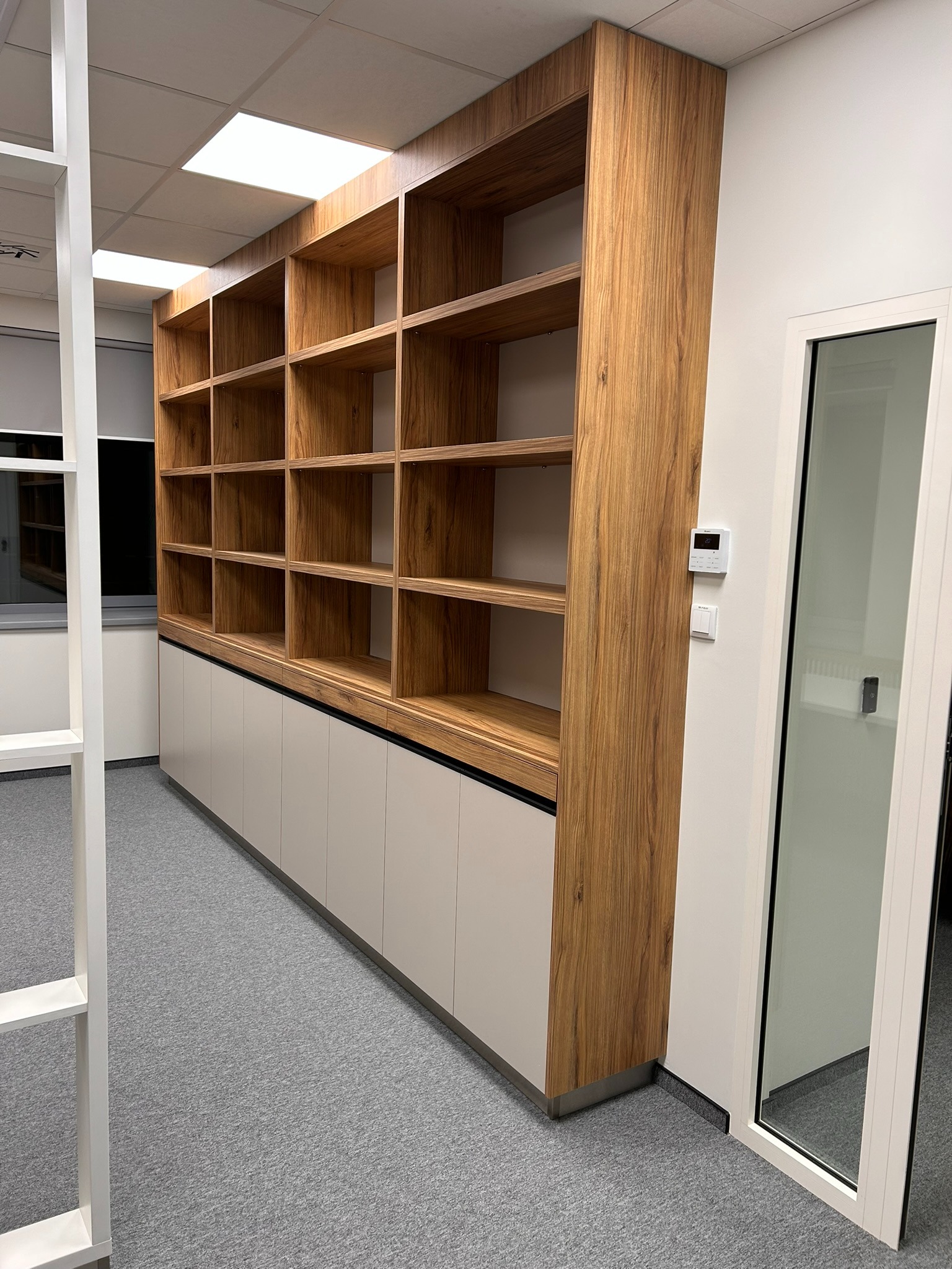 Drewniany regał z szafkami w biurze, widok z perspektywy. Naturalne wybarwienie drewna, minimalistyczne szafki, szara wykładzina. Nowoczesne oświetlenie sufitowe.