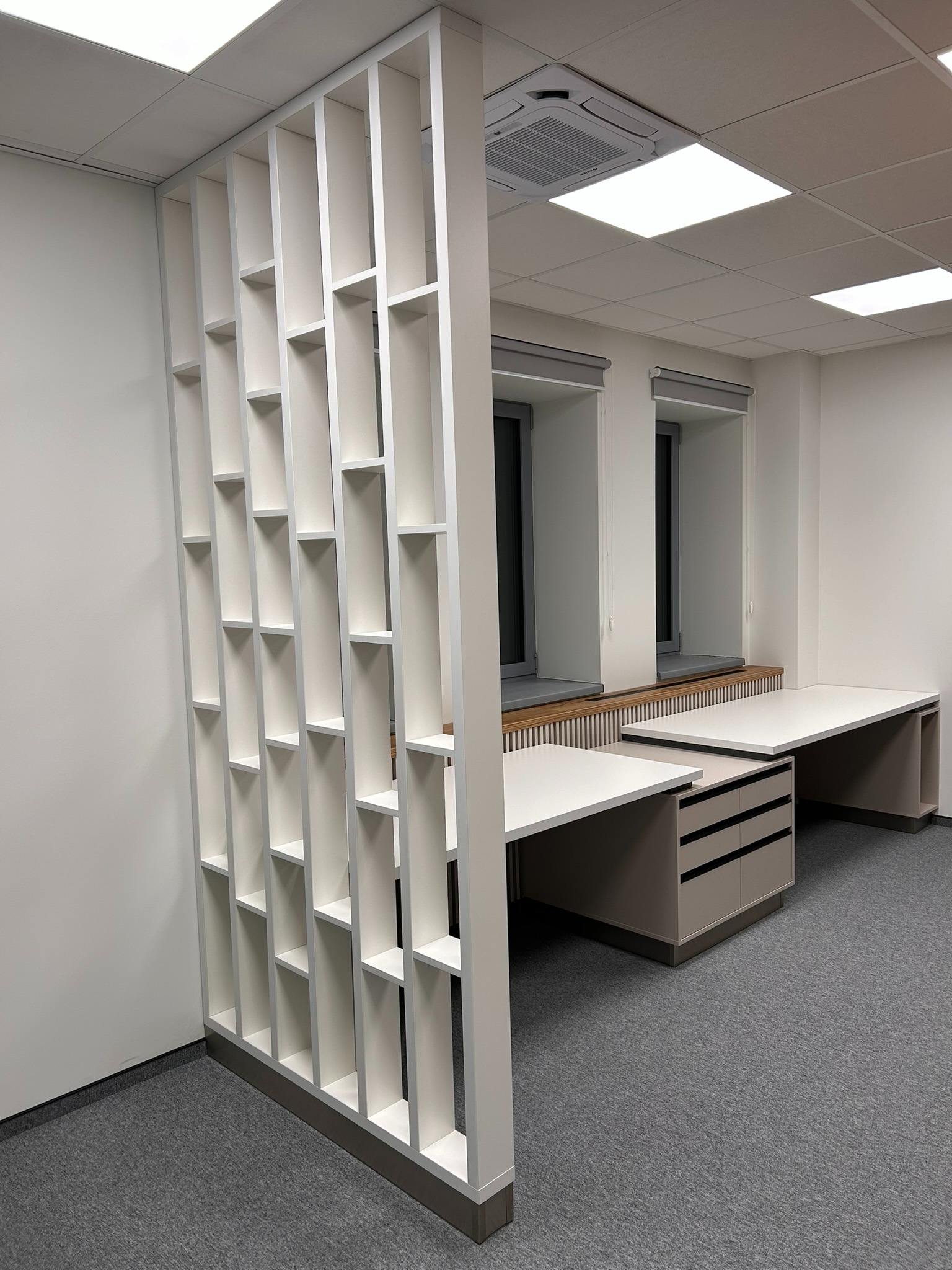 Biała, ażurowa ścianka działowa z MDF oddzielająca przestrzeń biurową z dwoma biurkami w neutralnej kolorystyce. Minimalistyczny design, szara wykładzina.