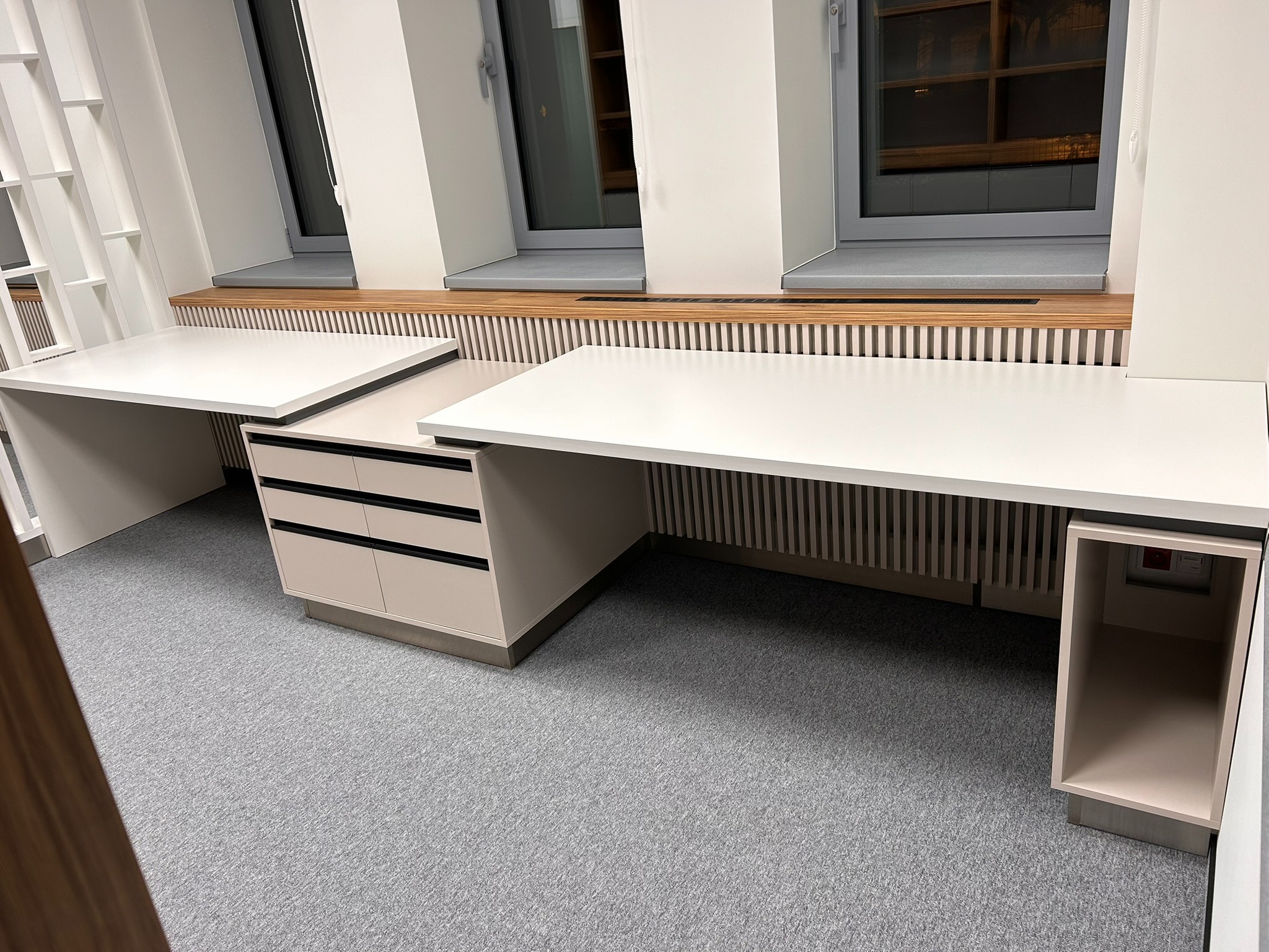 Dwa białe biurka z szufladami i szafką, ustawione pod oknami z drewnianymi panelami dekoracyjnymi. Szara wykładzina podłogowa. Minimalistyczny design.