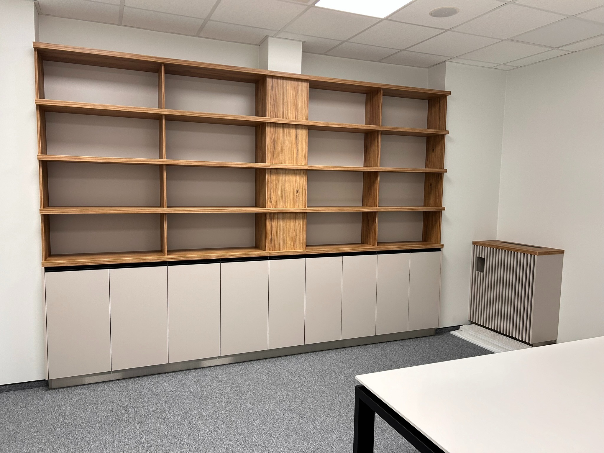 Regał z drewnianymi półkami i beżowymi szafkami dolnymi, uzupełniony grzejnikiem z lamelami. Minimalistyczny design, szara wykładzina podłogowa.