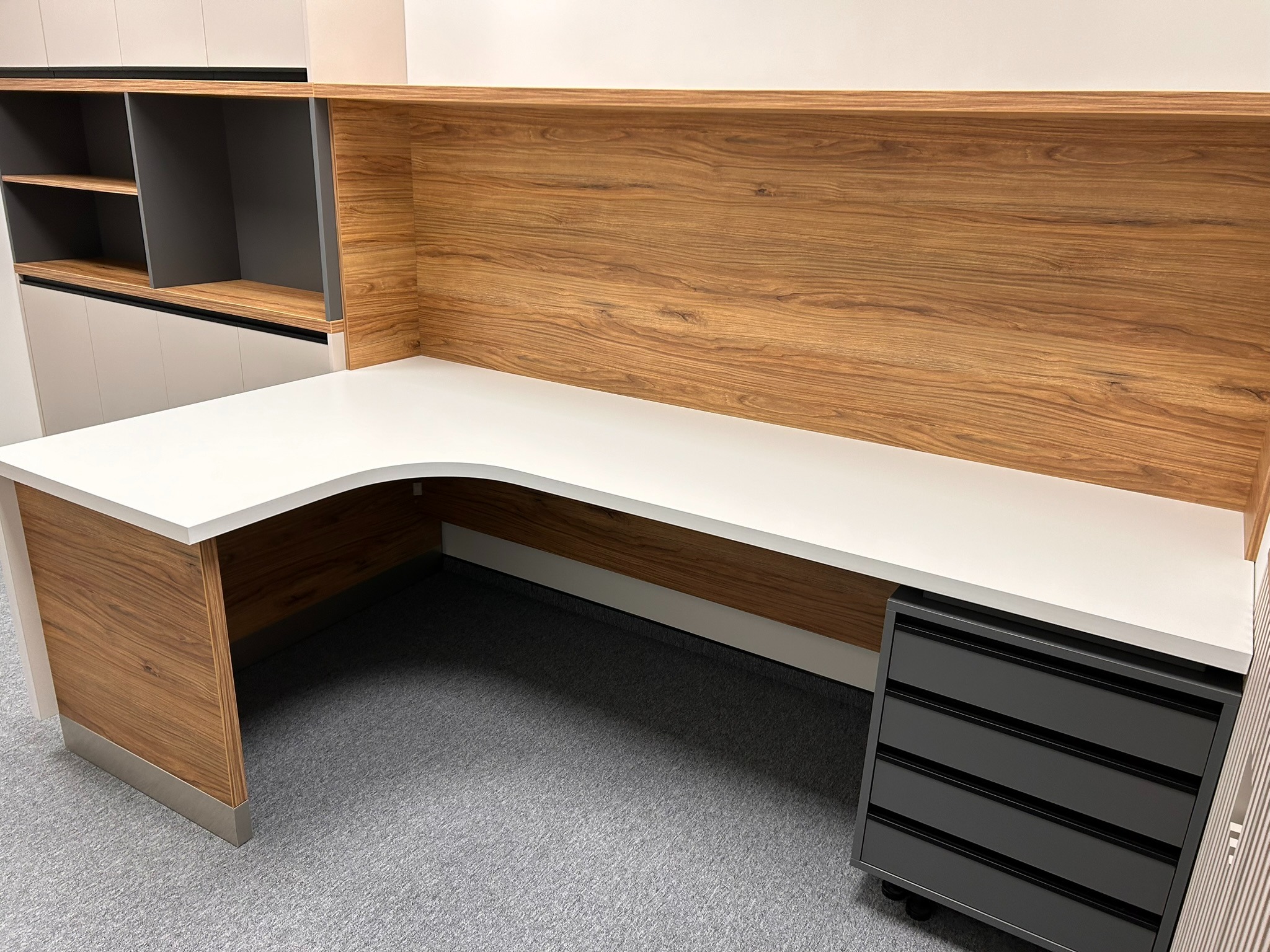 Narożne biurko z blatem w bieli, drewnianym panelem i szarą szafką na kółkach. Widoczne półki nad blatem. Nowoczesny design, jasne wnętrze.