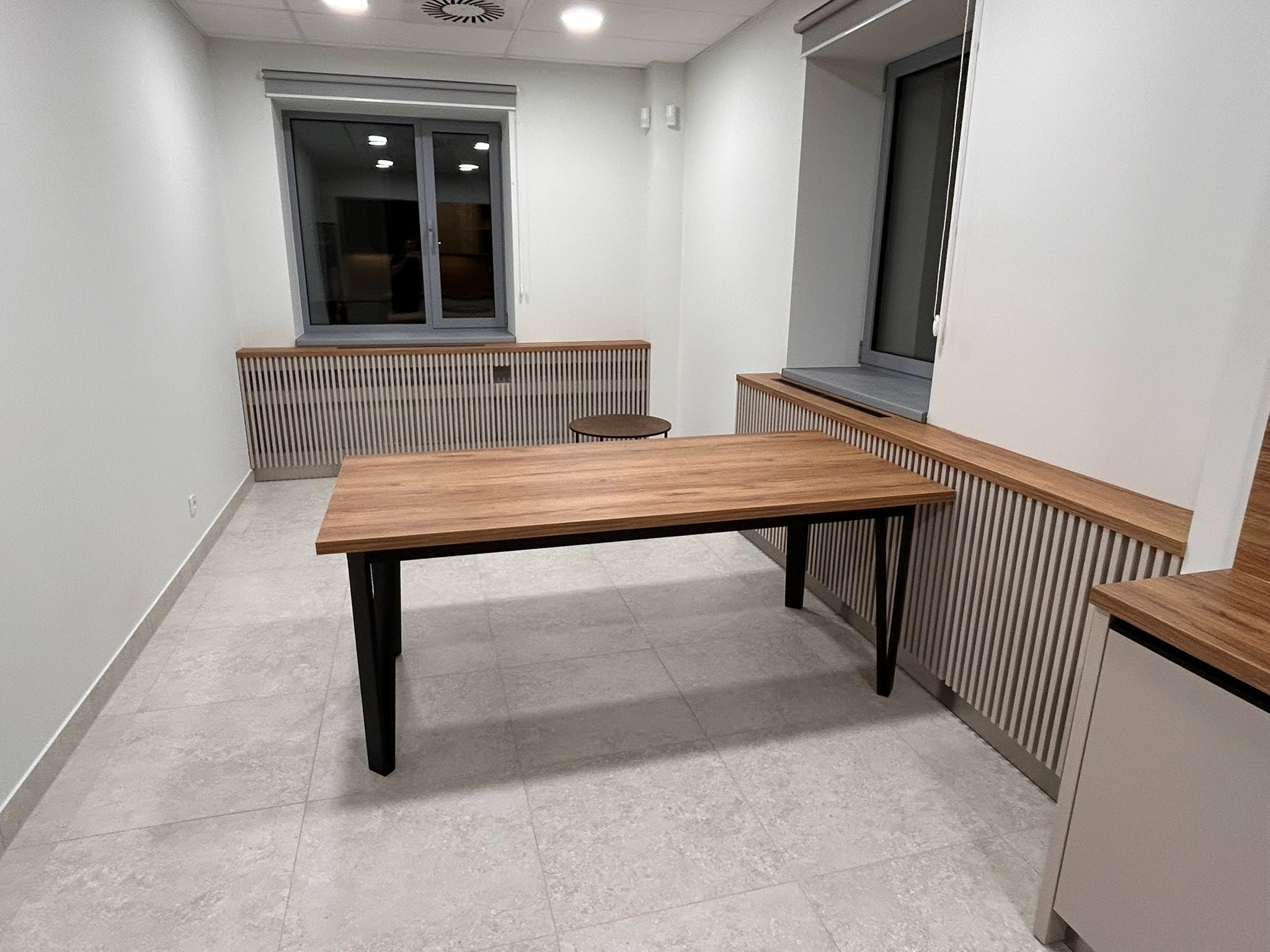 Stół z drewnianym blatem i czarnymi nogami w minimalistycznym wnętrzu z grzejnikami zabudowanymi w drewniane panele. Jasne płytki podłogowe i neutralna kolorystyka ścian.