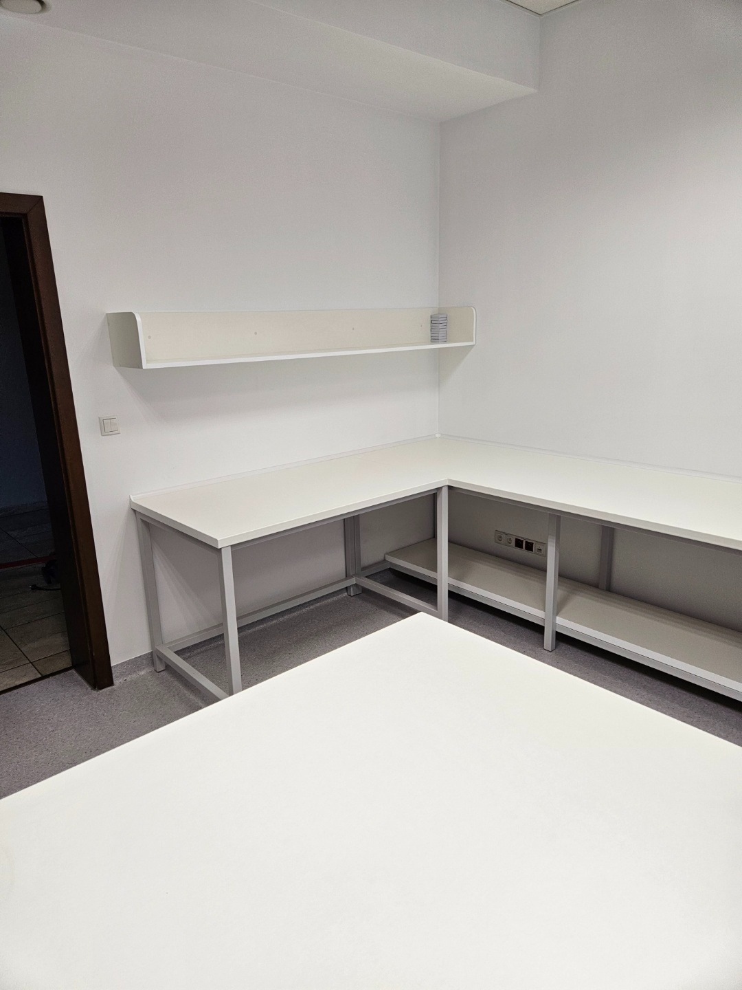 Białe biurko narożne z metalowym stelażem w minimalistycznym wnętrzu. Nad biurkiem półka z drobnymi przedmiotami. Widoczny fragment podłogi i drzwi.