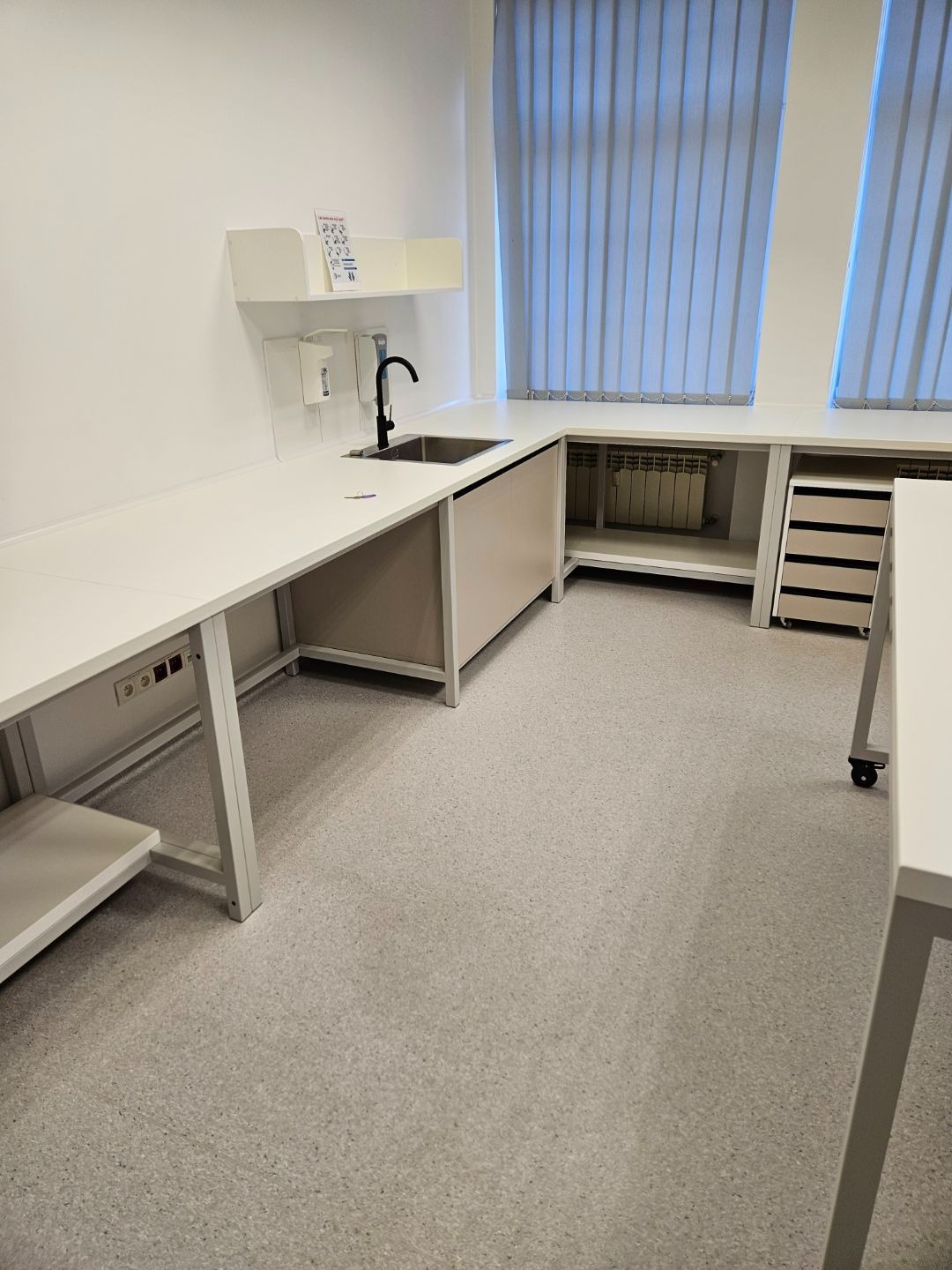 Jasna, nowoczesna zabudowa kuchenna z blatem roboczym, zlewem i szafkami w neutralnej kolorystyce. Widoczne okna z żaluzjami pionowymi i grzejnik.