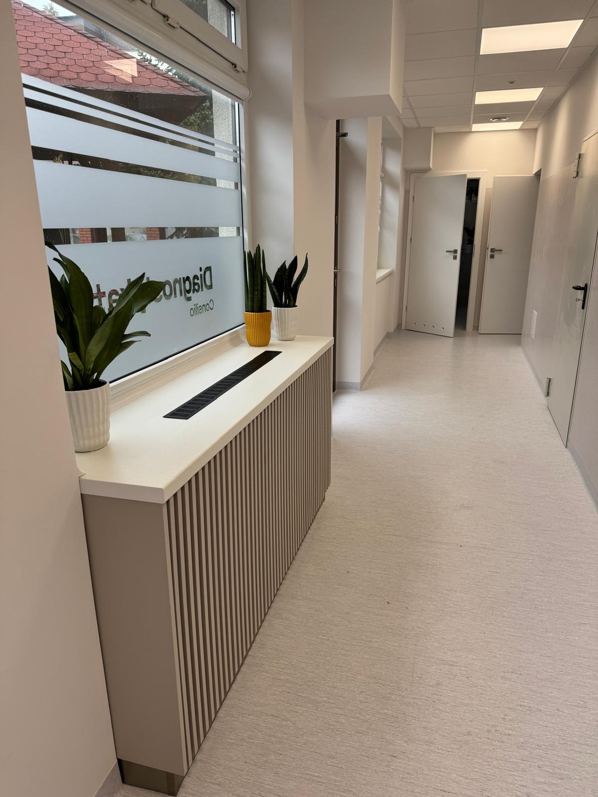 Biało-beżowa zabudowa grzejnika z pionowymi listwami, zwieńczona blatem z doniczkami, w jasnym korytarzu biurowym z oknem z folią dekoracyjną.