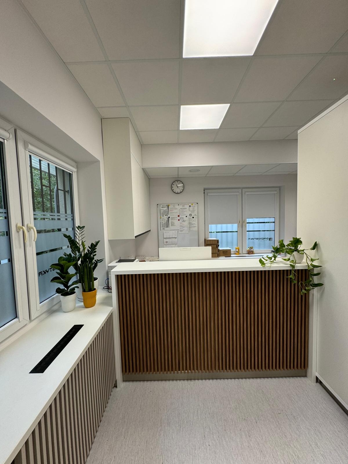 Nowoczesna recepcja z drewnianymi lamelami, jasnym blatem i roślinami doniczkowymi przy oknie. Minimalistyczny design, neutralna kolorystyka, widok na zewnątrz.