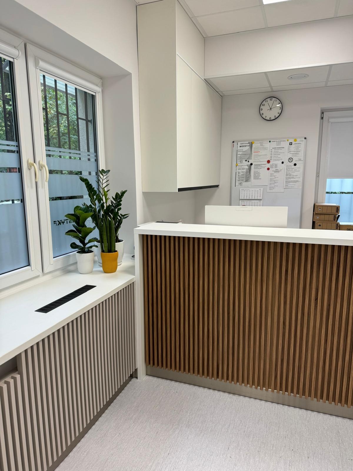 Nowoczesna recepcja z drewnianymi lamelami, biały blat, zabudowa okna z lamelami w odcieniach szarości, rośliny doniczkowe, minimalistyczny design, jasne wnętrze.