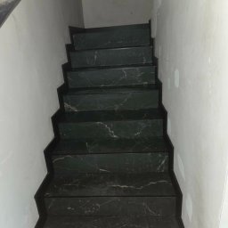 Jakub Krzywański - Wykończone schody wewnętrzne z ciemnego marmuru z widocznym wzorem żyłkowania, prowadzące w górę wzdłuż białej ściany. Widok z dołu na górę.