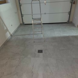 Jakub Krzywański - Wykończona podłoga z szarych, marmurkowych płytek w pomieszczeniu z białą bramą garażową i drabinką. Widoczny odpływ liniowy.