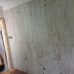 Jakub Krzywański - Ściana wyłożona beżowymi płytkami imitującymi kamień, z widocznymi śladami po montażu i wystającymi przewodami. W tle ościeżnica drzwiowa i płyta karton-gips.