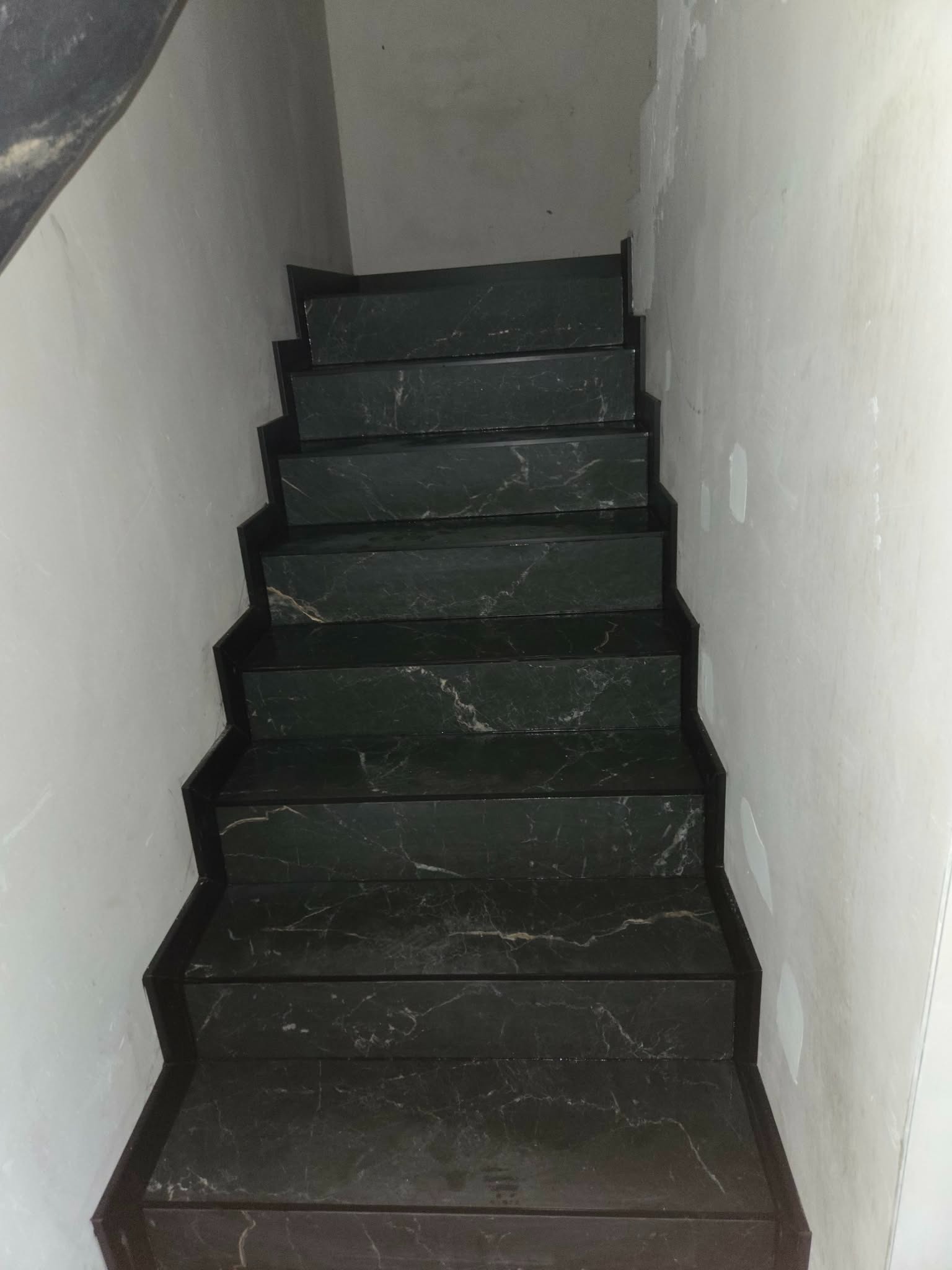 Wykończone schody wewnętrzne z ciemnego marmuru z widocznym wzorem żyłkowania, prowadzące w górę wzdłuż białej ściany. Widok z dołu na górę.