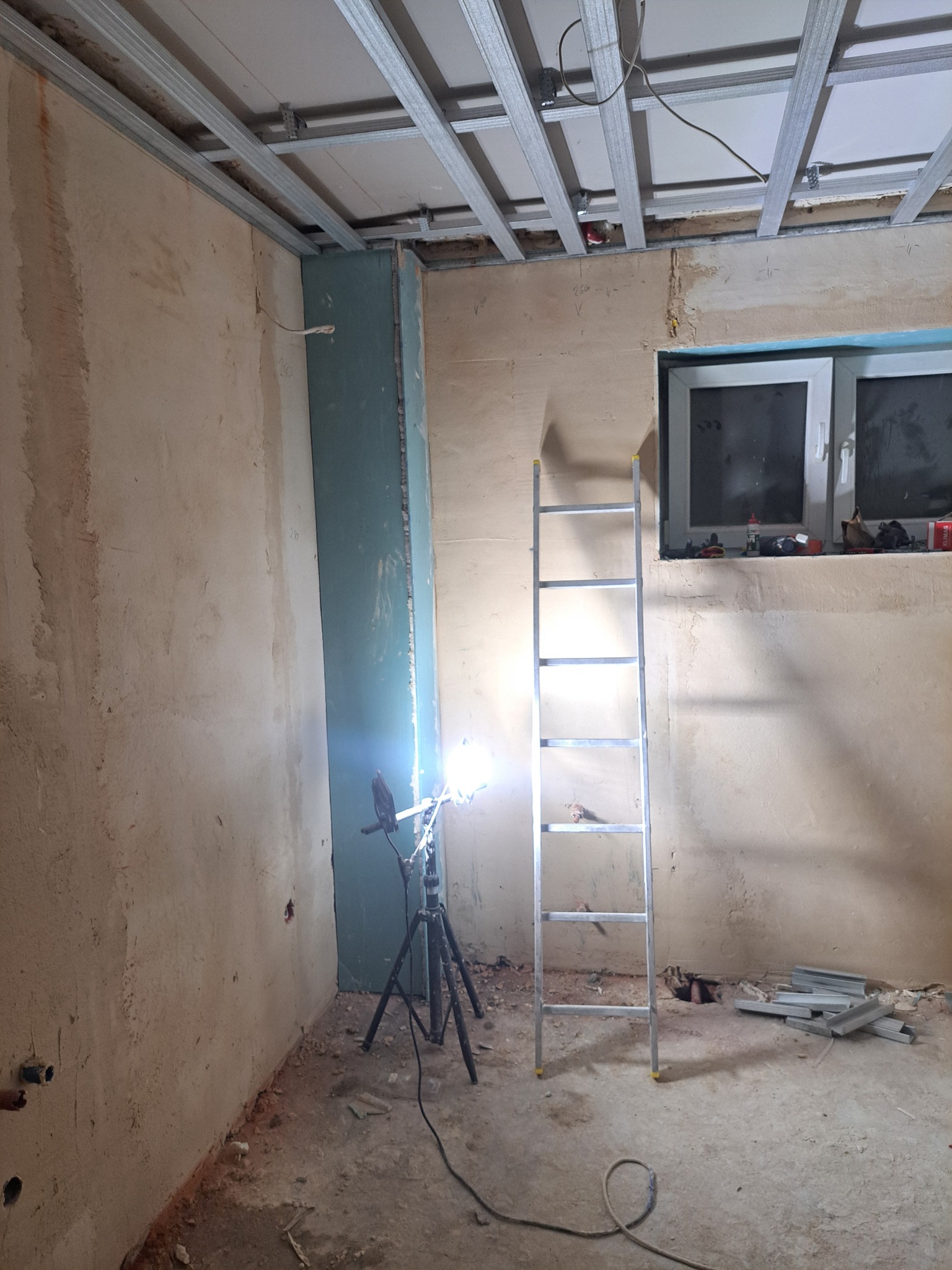 Wnętrze w trakcie montażu sufitu podwieszanego z metalowej konstrukcji. Widoczna drabina, lampa na statywie i fragment ściany z płytą karton-gips.