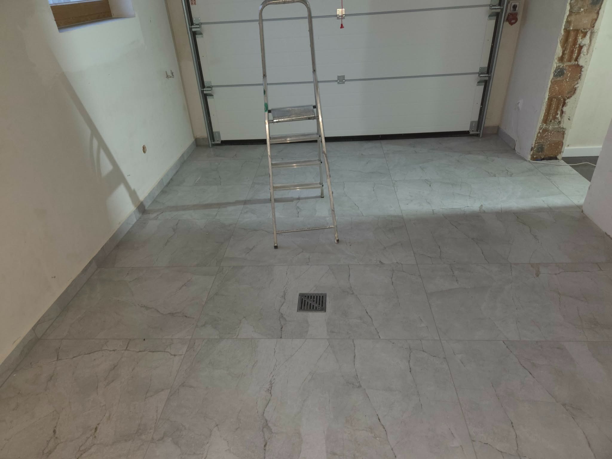 Wykończona podłoga z szarych, marmurkowych płytek w pomieszczeniu z białą bramą garażową i drabinką. Widoczny odpływ liniowy.