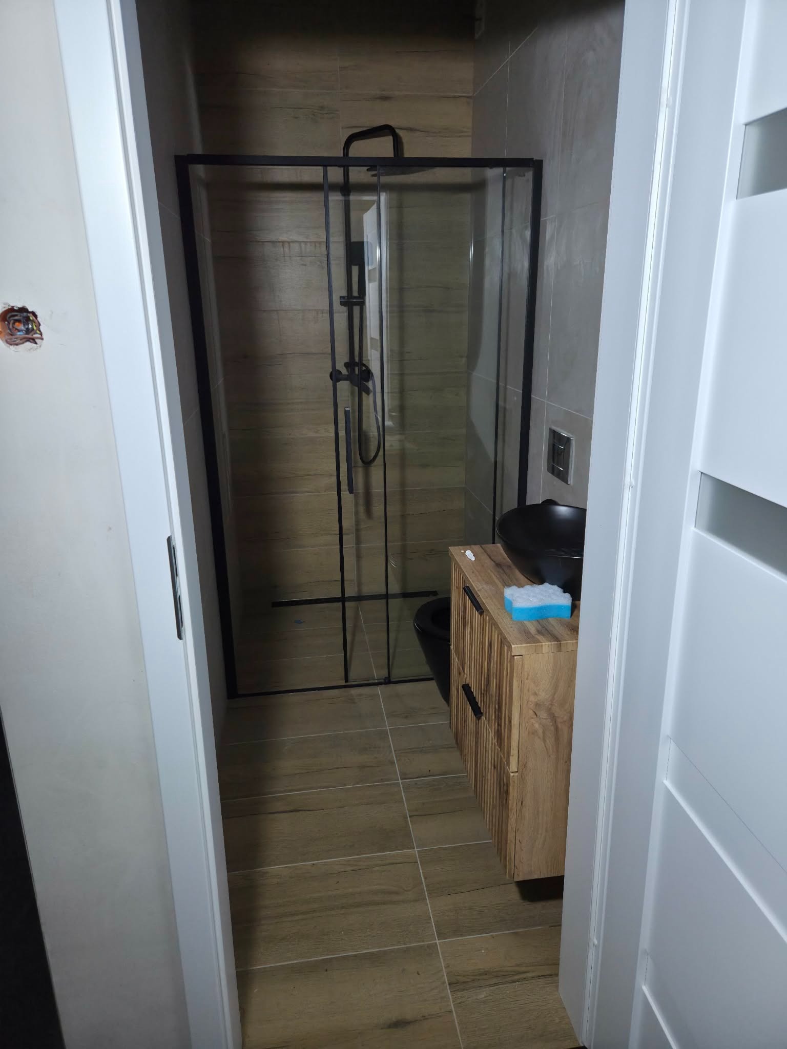 Nowoczesna łazienka po remoncie: kabina prysznicowa z czarną ramą, drewniana szafka z czarną umywalką, płytki imitujące drewno na podłodze i ścianie. Widok zza otwartych drzwi.