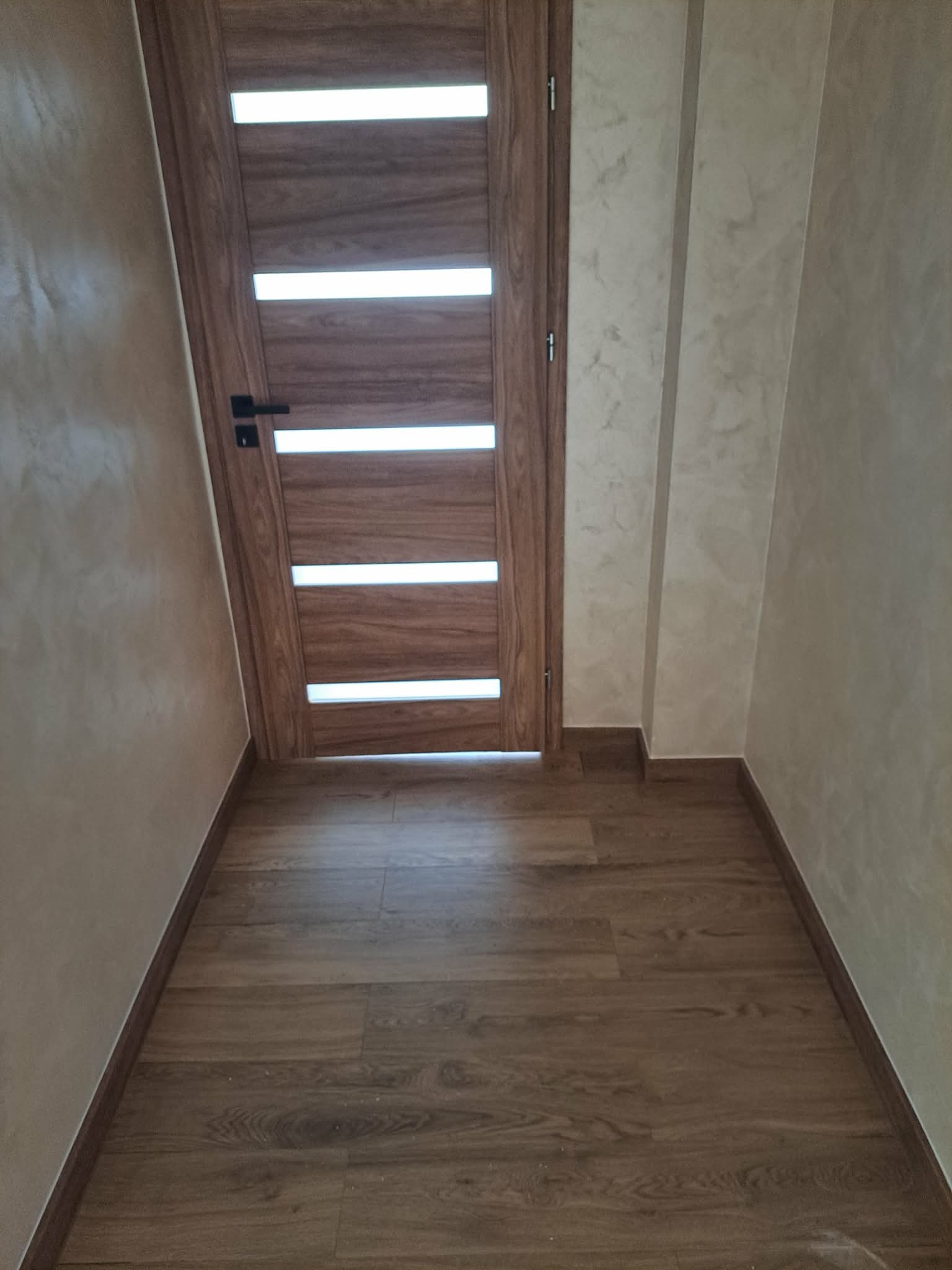 Nowoczesne wnętrze: drewniana podłoga, drzwi z przeszkleniami i ściany w odcieniach beżu z delikatną fakturą. Minimalistyczny design, ciepłe kolory, wykończone detale.