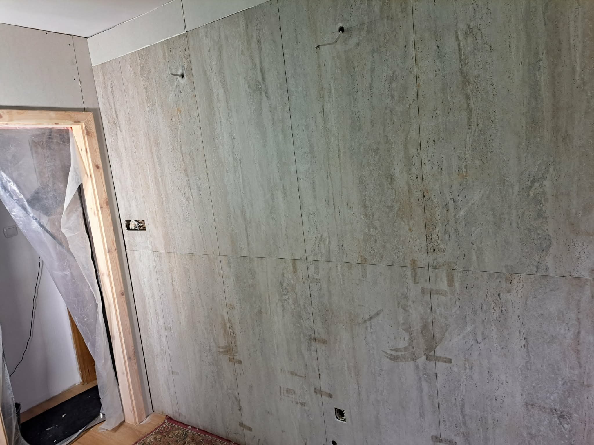 Ściana wyłożona beżowymi płytkami imitującymi kamień, z widocznymi śladami po montażu i wystającymi przewodami. W tle ościeżnica drzwiowa i płyta karton-gips.