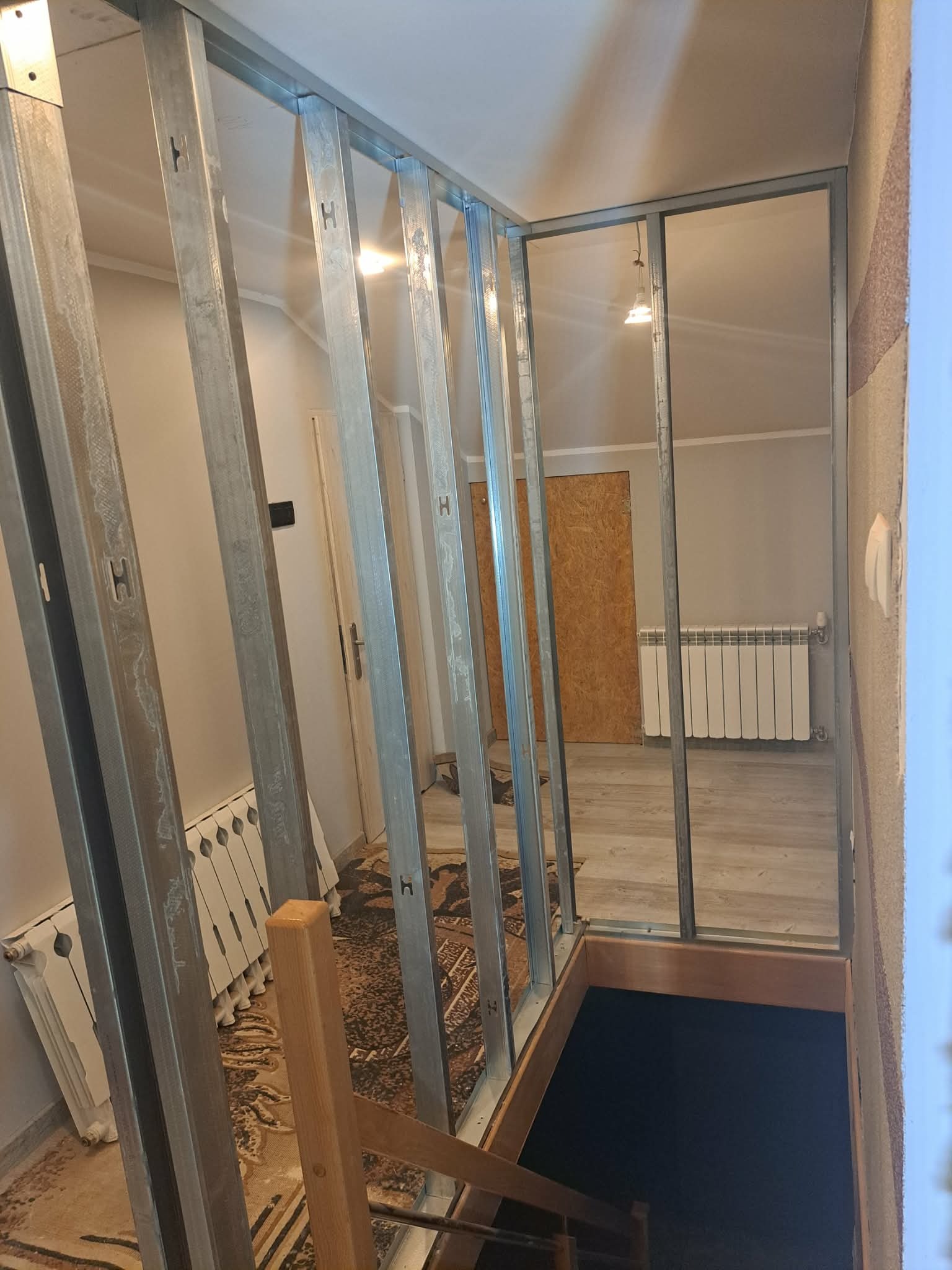 Metalowa konstrukcja ścianki działowej z profili, widok z góry schodów. W tle widoczny grzejnik i płyta OSB. Etap prac wykończeniowych w pomieszczeniu.