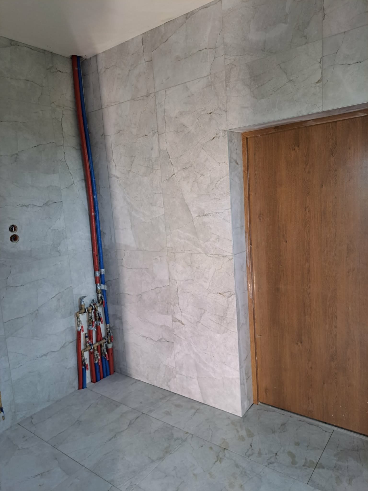 Wykończona łazienka z marmurowymi płytkami, widoczne rury instalacji wodnej w kolorze niebieskim i czerwonym oraz drewniane drzwi. Minimalistyczny design.