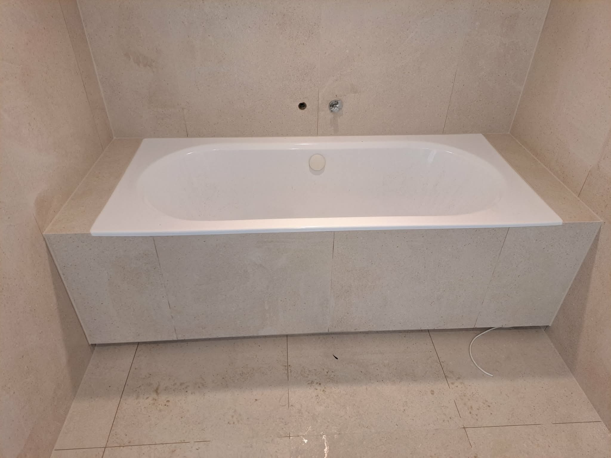 Biała wanna obudowana beżowymi płytkami w łazience. Widoczne fugi i otwór na baterię. Minimalistyczny design, ciepłe odcienie.