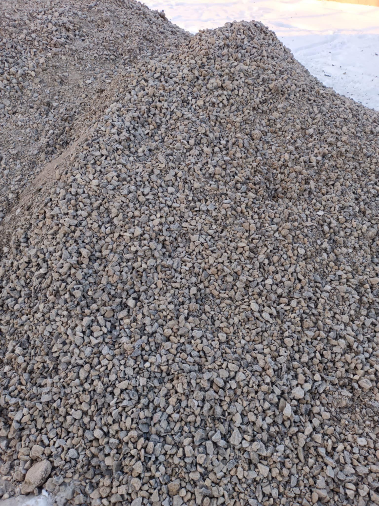 Kupa szarego kruszywa o nieregularnych kształtach, z widocznym śniegiem w tle, sugerująca materiał budowlany lub drogowy.