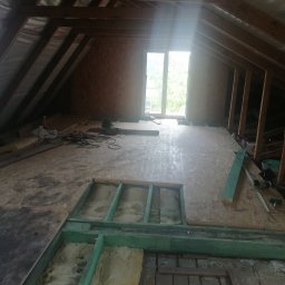 Poddasze w trakcie adaptacji: widoczne ocieplenie wełną mineralną między legarami, częściowo położona płyta OSB. W tle okno dachowe i drewniana konstrukcja dachu.