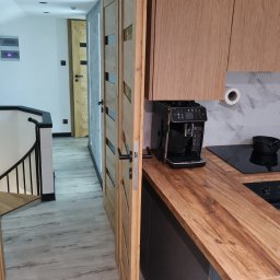 Kamil Melaniuk - Nowoczesna kuchnia z drewnianym blatem i frontami, widok na otwarte drzwi z przeszkleniami i schody. Minimalistyczny design, jasne kolory, ekspres do kawy na blacie.