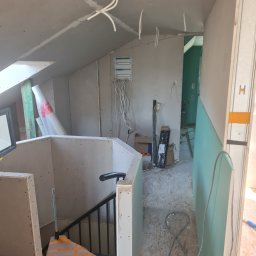 Kamil Melaniuk - Skośne poddasze w trakcie wykańczania płytami gipsowo-kartonowymi, widoczne kable elektryczne, fragment schodów i materiały budowlane. Prace wykończeniowe w surowym stanie.