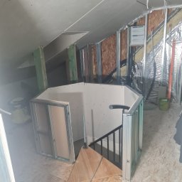 Adaptacja poddasza: zabudowa schodów płytą gipsowo-kartonową na metalowym stelażu. Widoczne elementy konstrukcyjne i izolacja. Prace w toku, sufit z płyt.