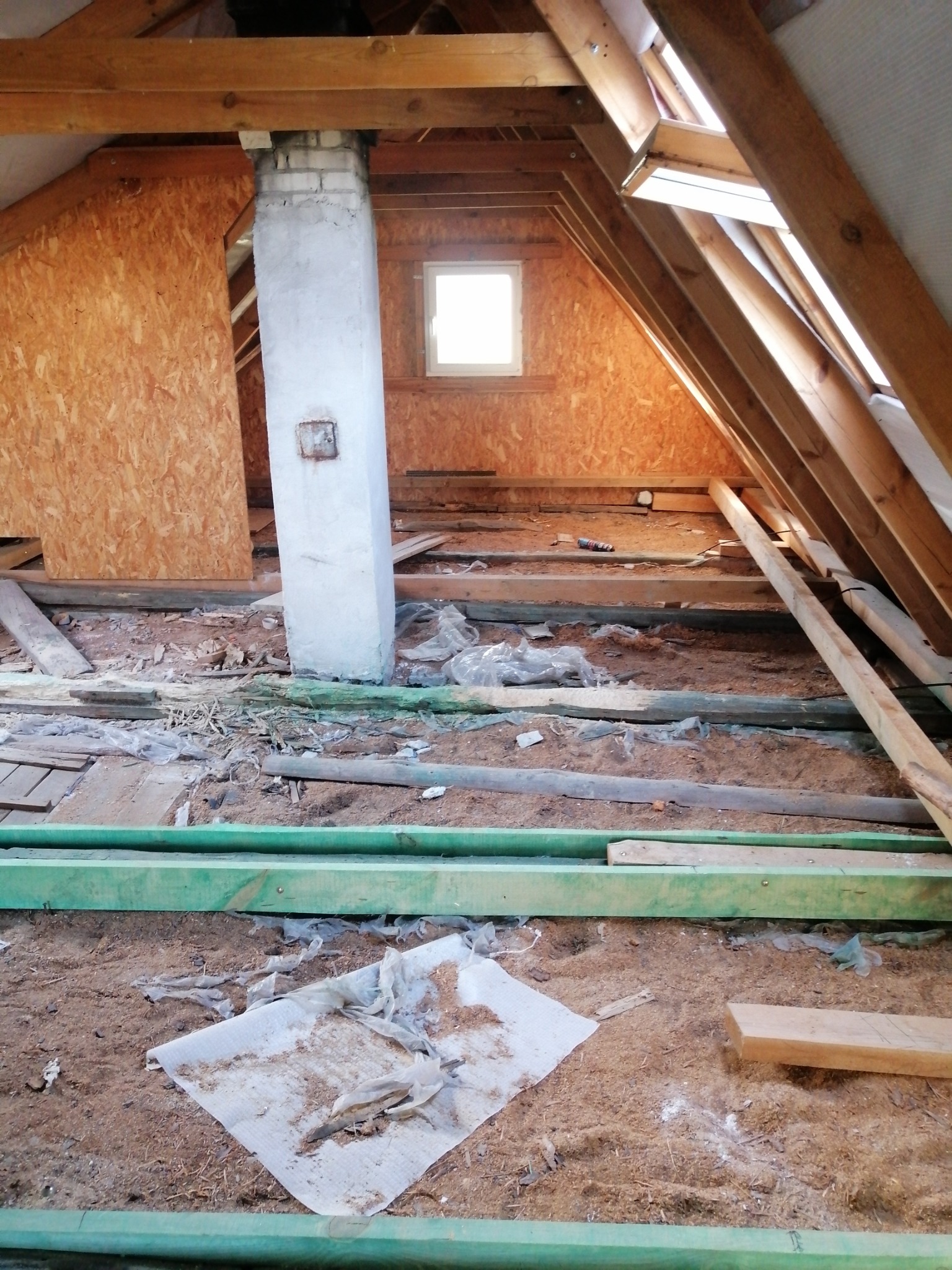 Poddasze w trakcie adaptacji: widoczne drewniane belki, fragment komina, okno dachowe i płyty OSB. Podłoga pokryta trocinami i folią, przygotowana do dalszych prac.