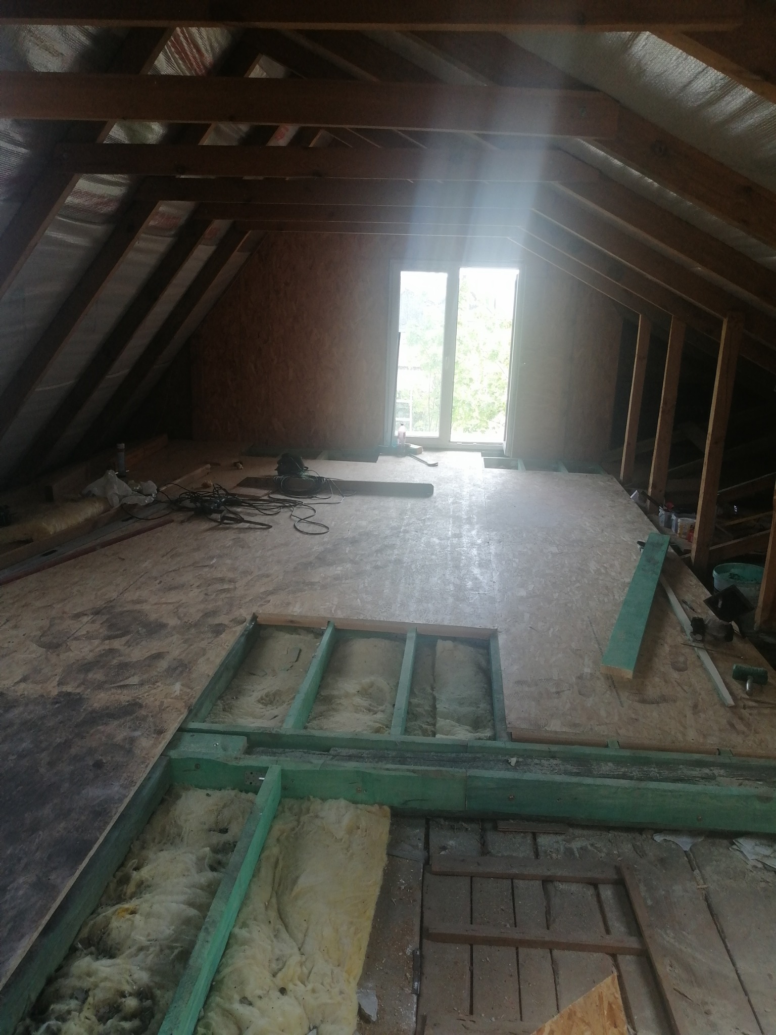 Poddasze w trakcie adaptacji: widoczne ocieplenie wełną mineralną między legarami, częściowo położona płyta OSB. W tle okno dachowe i drewniana konstrukcja dachu.