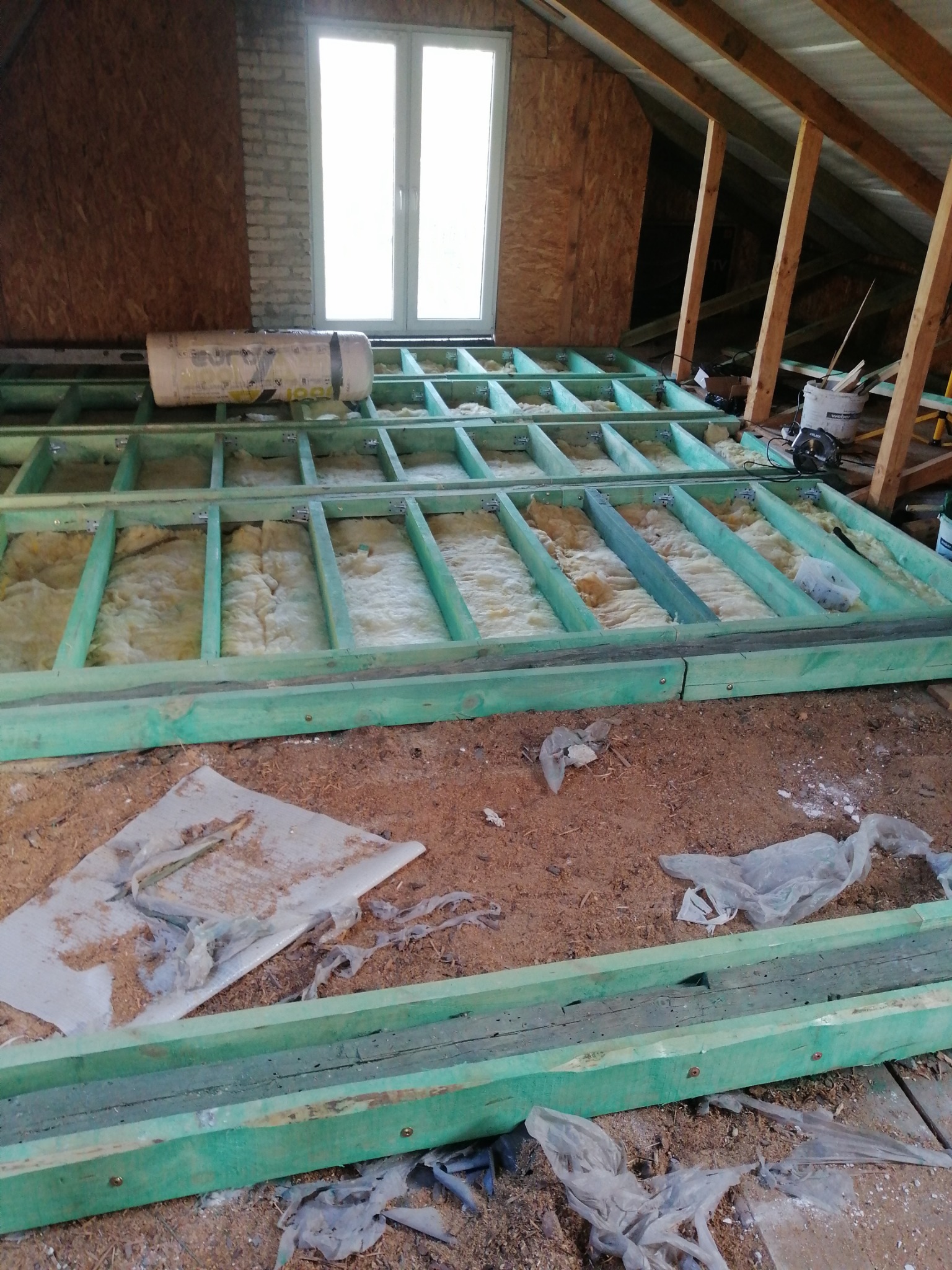 Ocieplenie poddasza: drewniana konstrukcja z wełną mineralną, widoczne elementy mocujące, okno w tle. Etap prac wykończeniowych na poddaszu.