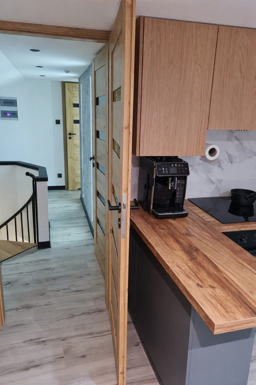 Nowoczesna kuchnia z drewnianym blatem i frontami, widok na otwarte drzwi z przeszkleniami i schody. Minimalistyczny design, jasne kolory, ekspres do kawy na blacie.