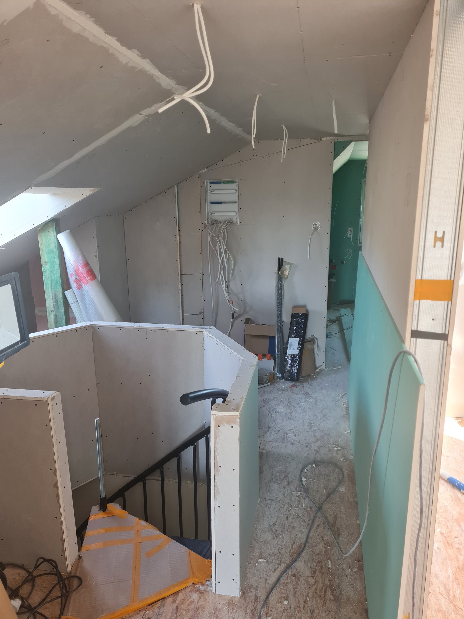 Skośne poddasze w trakcie wykańczania płytami gipsowo-kartonowymi, widoczne kable elektryczne, fragment schodów i materiały budowlane. Prace wykończeniowe w surowym stanie.