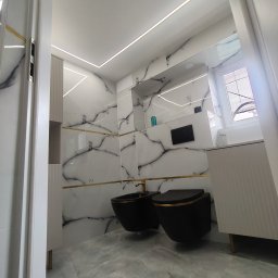 Gres Pro - Nowoczesna łazienka z czarną armaturą i marmurowymi płytkami, oświetlenie LED. Ujęcie zza framugi drzwi, podkreślające głębię i wykończenie.