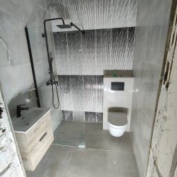 Gres Pro - Nowoczesna łazienka w szarościach z kabiną prysznicową typu walk-in, umywalką z szafką z jasnego drewna i podwieszaną toaletą. Ściany wyłożone płytkami z wzorem.