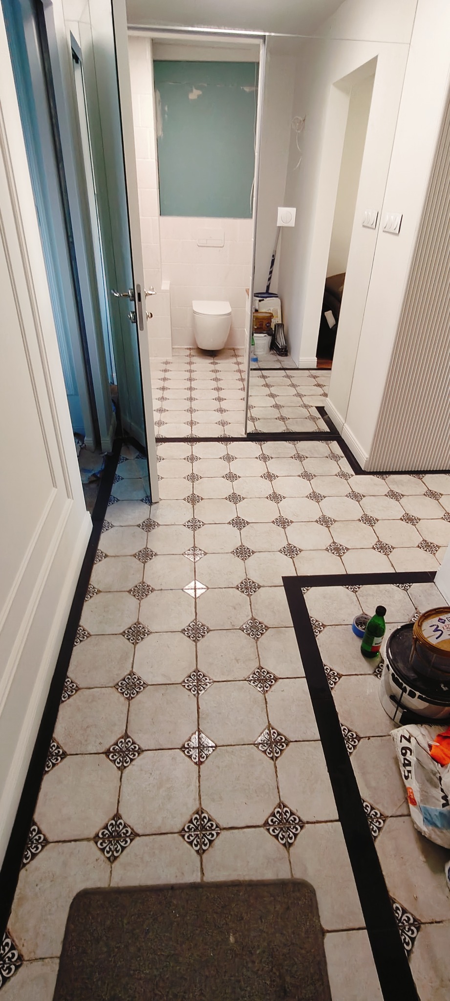 Ukończona podłoga w łazience z wzorzystymi płytkami i czarnymi listwami. Widoczne akcesoria malarskie i fragment toalety w tle. Ujęcie z perspektywy wejścia.