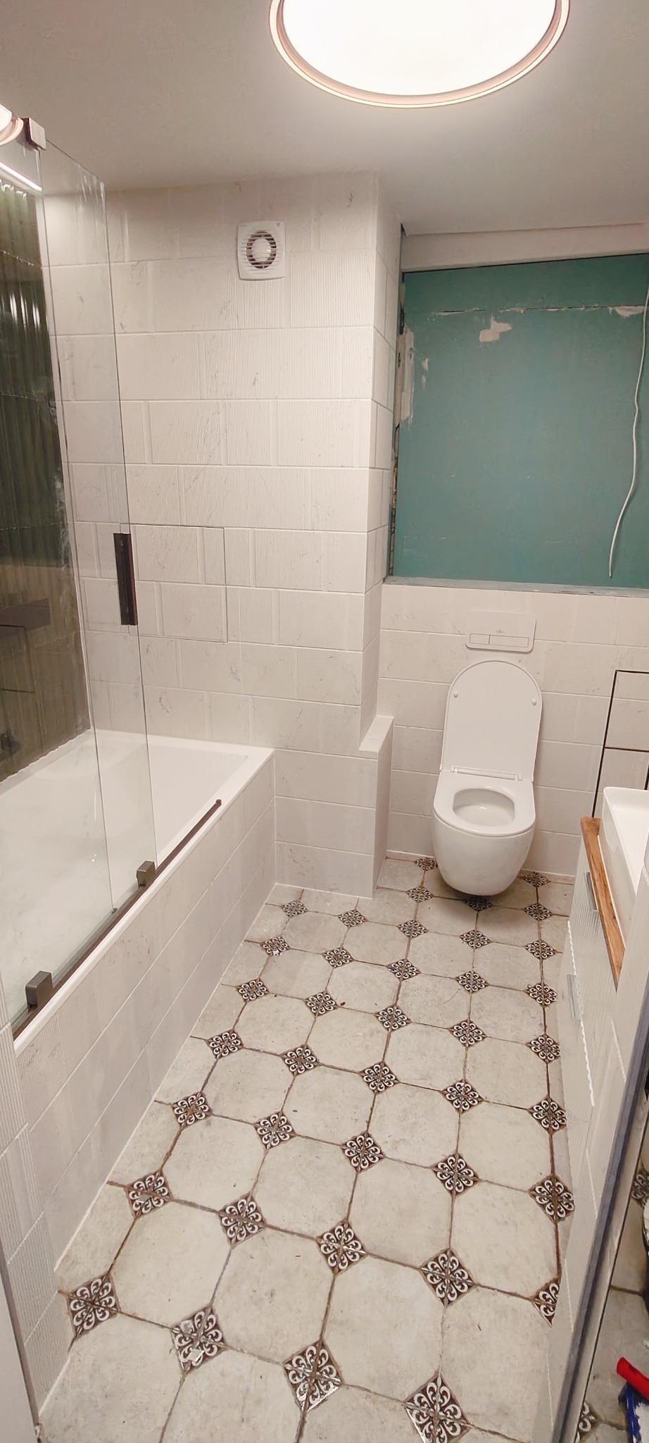 Wykończona łazienka z wanną za szklaną ścianką, toaletą i umywalką. Na podłodze płytki w stylu retro. Ściany wyłożone białymi, podłużnymi płytkami.