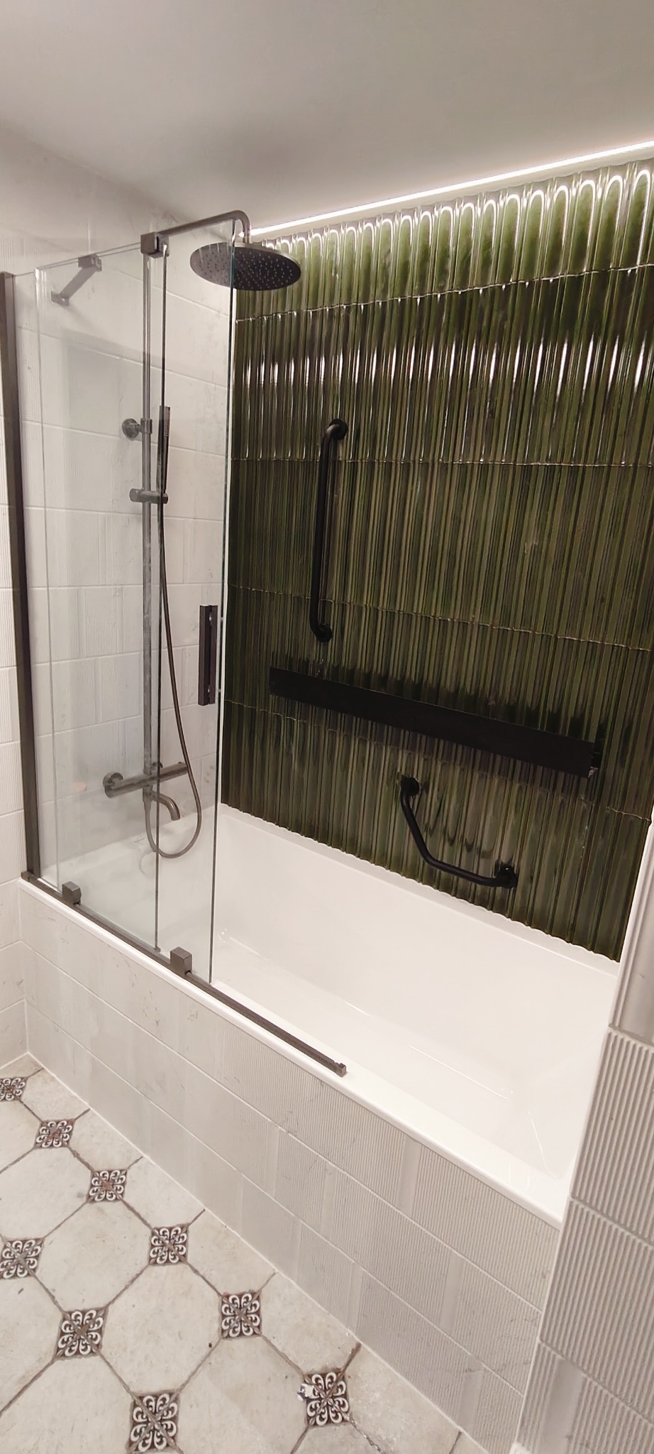 Nowoczesna łazienka z wanną i szklanym parawanem, zielone płytki na ścianie, wzorzysta podłoga. Minimalistyczny design z akcentami retro.