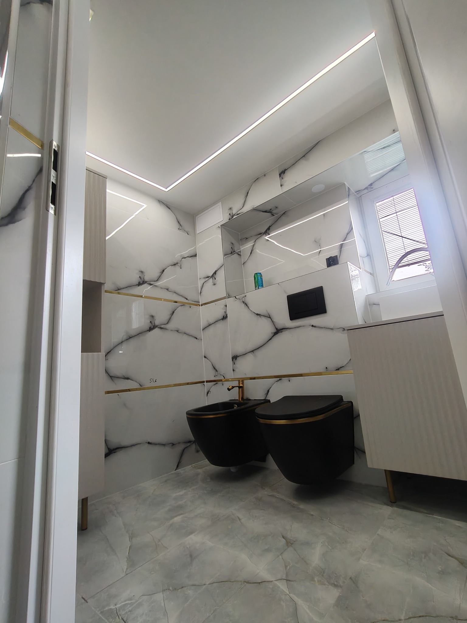 Nowoczesna łazienka z czarną armaturą i marmurowymi płytkami, oświetlenie LED. Ujęcie zza framugi drzwi, podkreślające głębię i wykończenie.