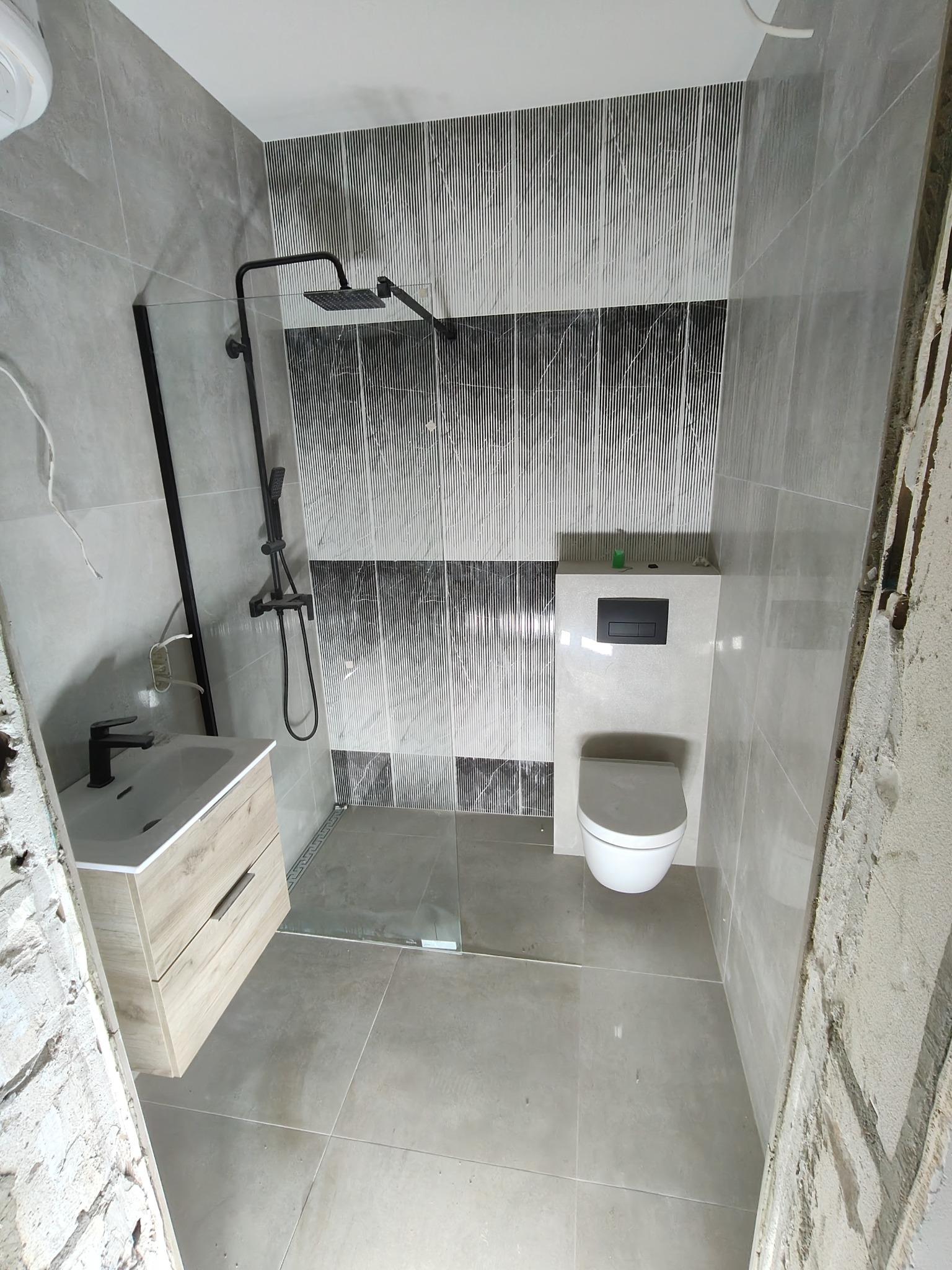 Nowoczesna łazienka w szarościach z kabiną prysznicową typu walk-in, umywalką z szafką z jasnego drewna i podwieszaną toaletą. Ściany wyłożone płytkami z wzorem.